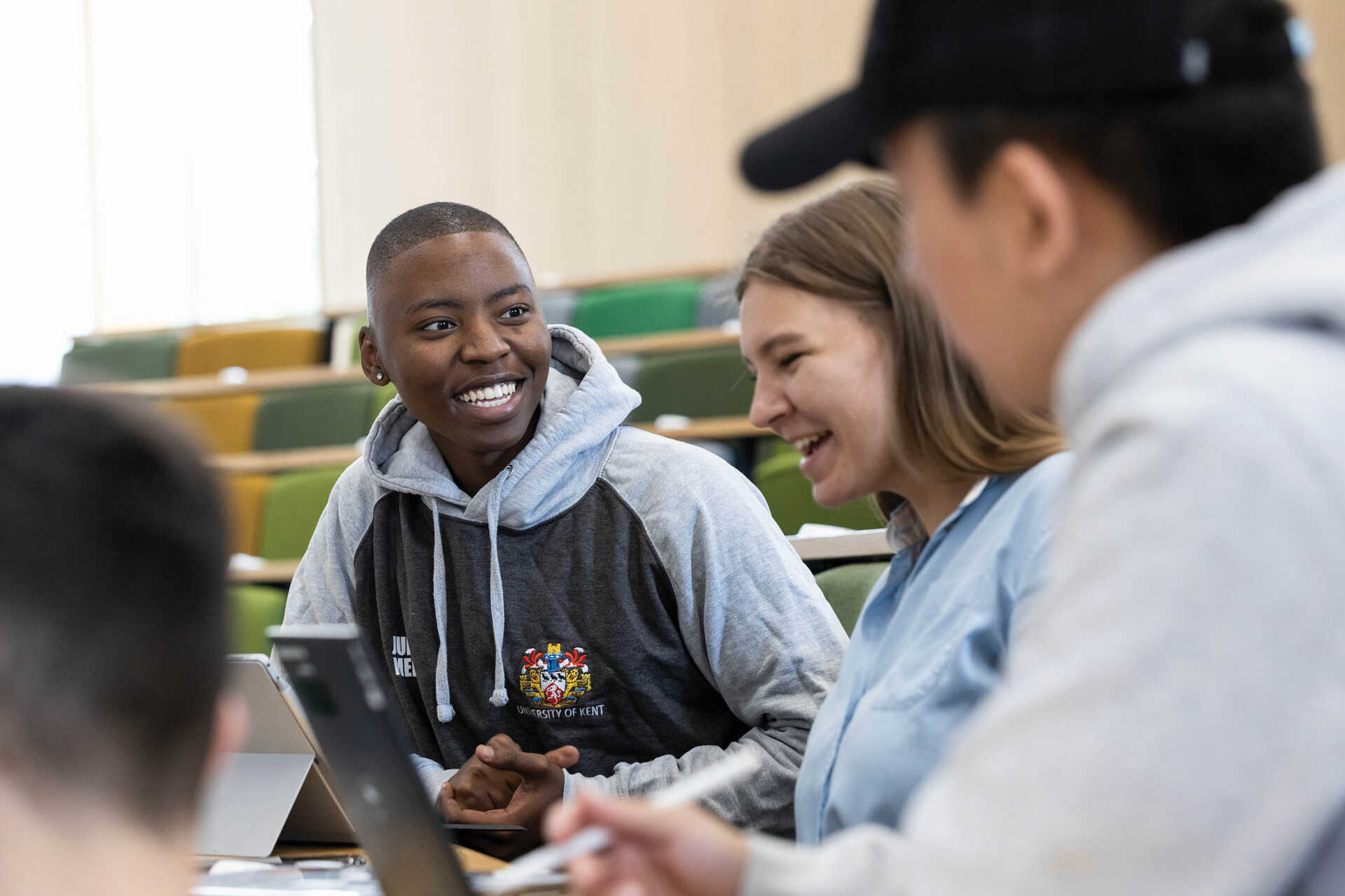 Students in a lecture theatre in conversation. One student is looking at a laptop. They are smiling.