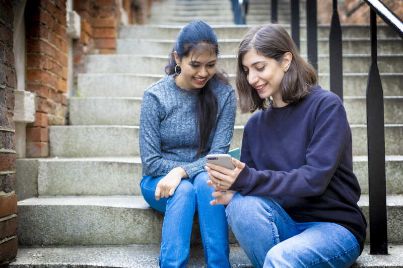 Two Kent students sitting on steps looking at a mobile phone together, smiling.